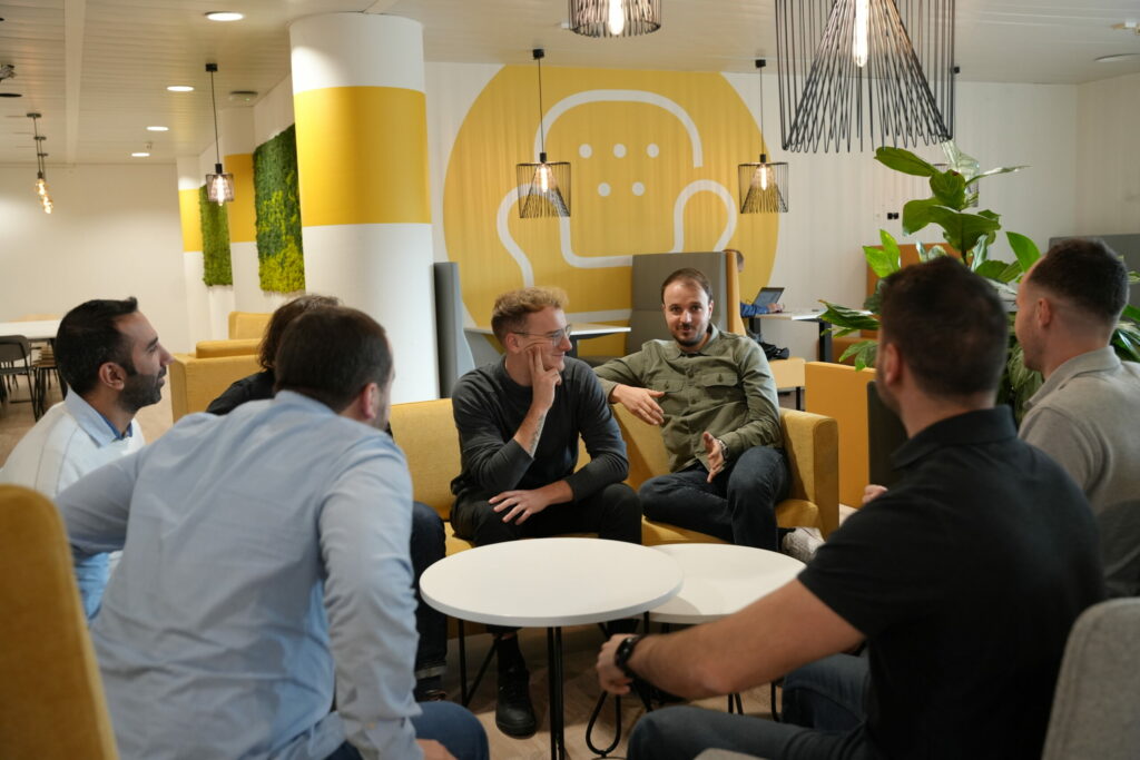 Un groupe de sept personnes sont assises autour de deux tables basses blanches dans un salon de bureau confortable et moderne aux murs jaunes et blancs. Elles discutent en toute émotion, sourient et rient, assises sur des canapés et des chaises jaunes.
