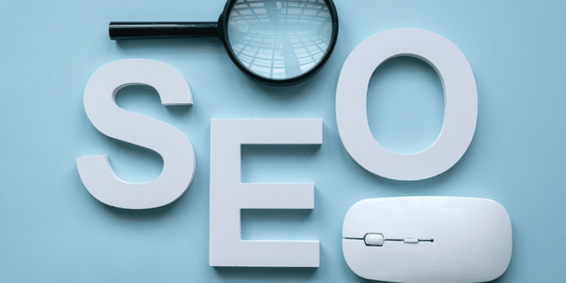 Lettres blanches formant le sigle SEO avec une loupe et une souris d’ordinateur sur fond bleu clair.