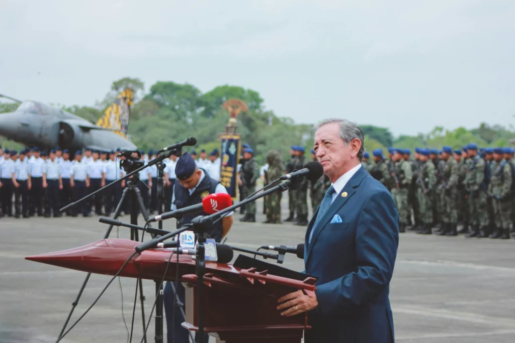 Un homme en costume prononce un discours devant une foule de militaires, avec un avion en arrière-plan.