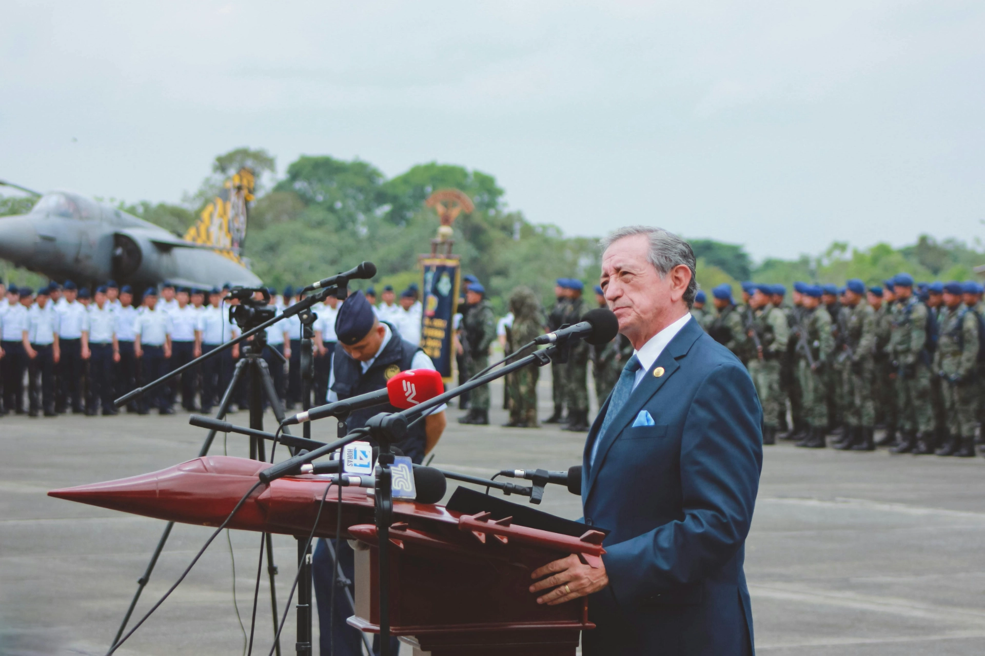 Un homme en costume prononce un discours devant une foule de militaires, avec un avion en arrière-plan.