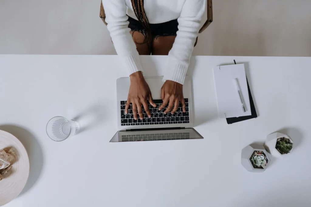 Vue de dessus d’une femme tapant sur un ordinateur portable posé sur un bureau blanc avec un carnet, un verre d’eau et deux petites plantes en pot.