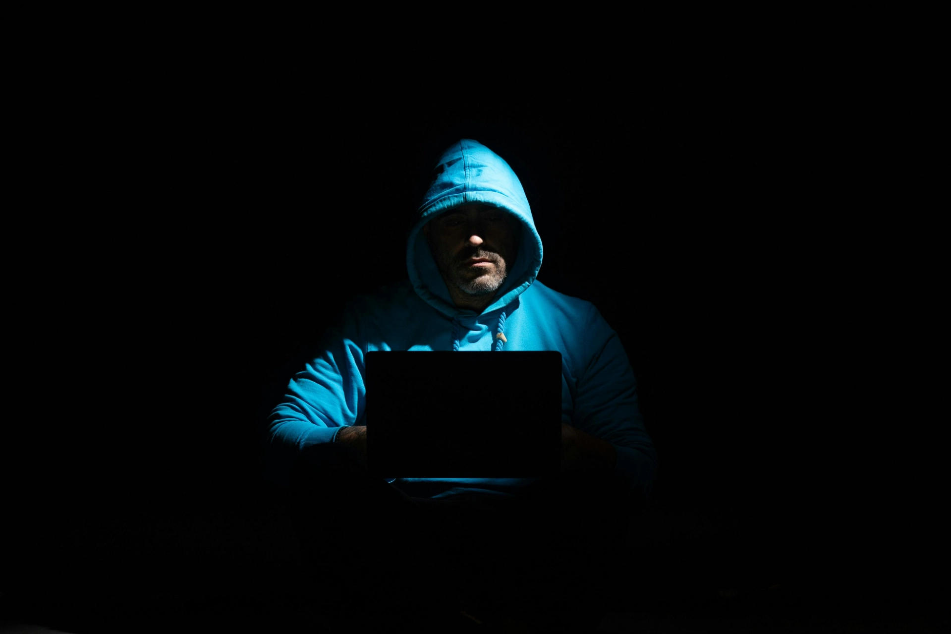 Homme en sweat à capuche bleu utilisant un ordinateur portable dans l’obscurité.