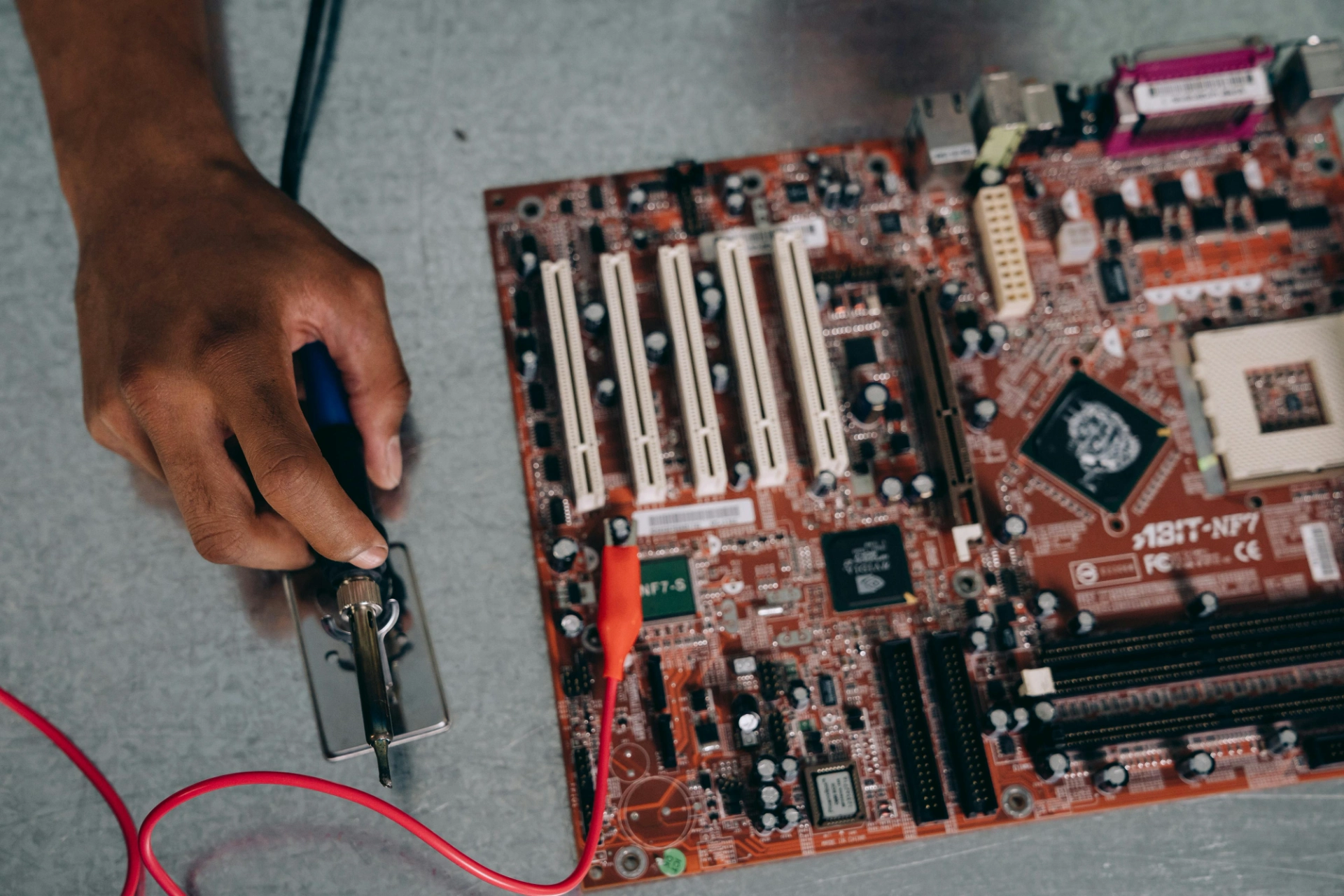 Main tenant un fer à souder près d’une carte mère électronique avec des fils connectés.