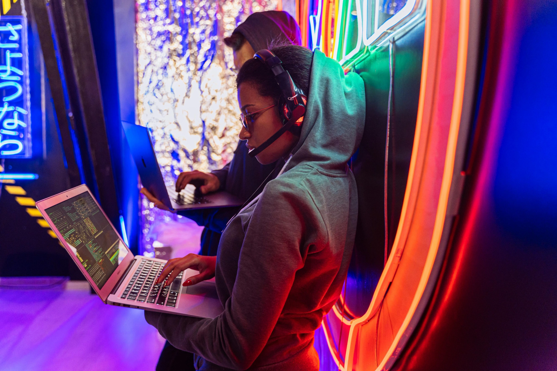 Femme en hoodie tapant sur un ordinateur portable avec des codes affichés à l’écran, accompagnée d’un collègue dans un environnement lumineux.