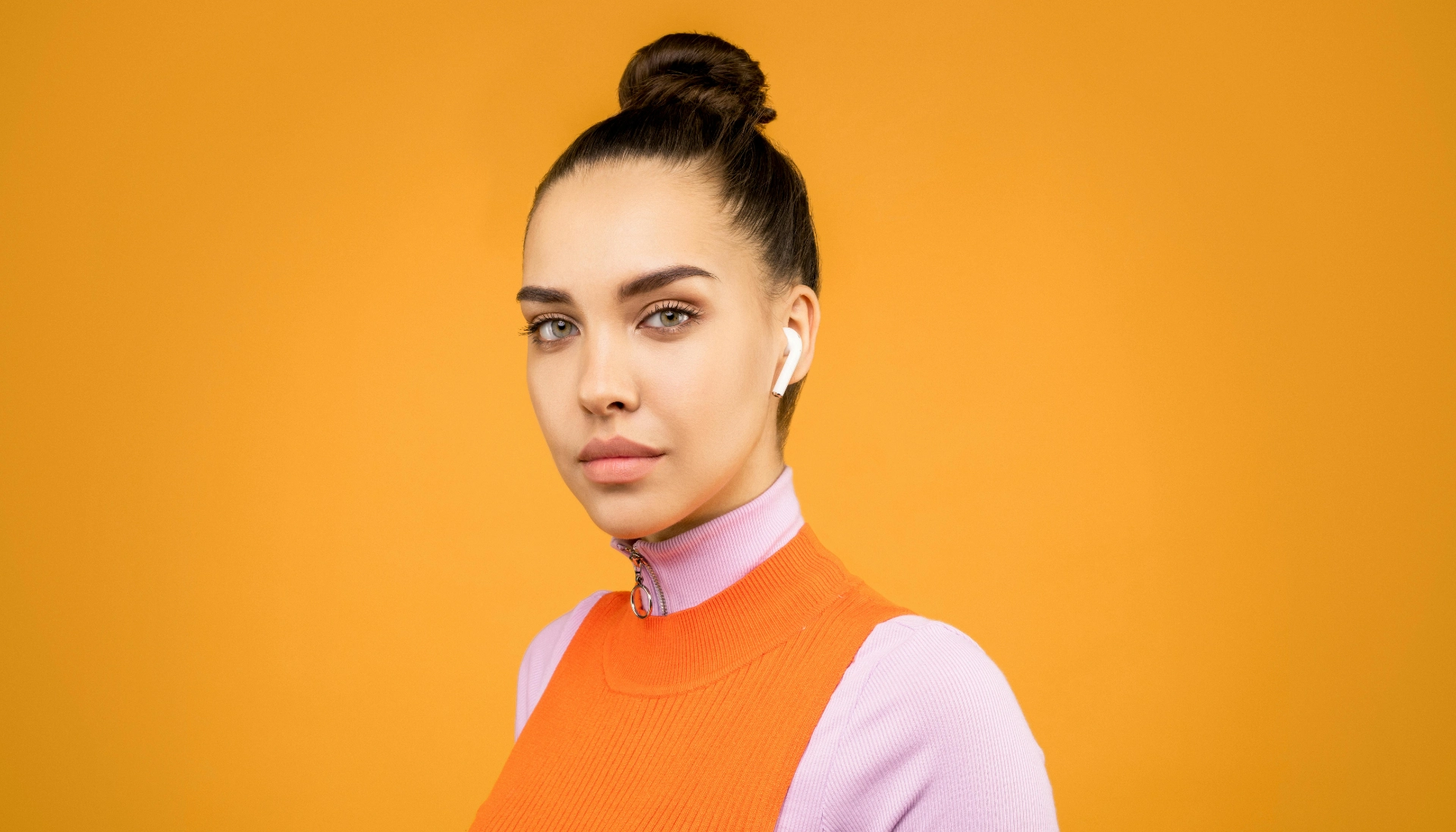 Une jeune femme portant des écouteurs sans fil, avec un pull orange et un fond jaune.