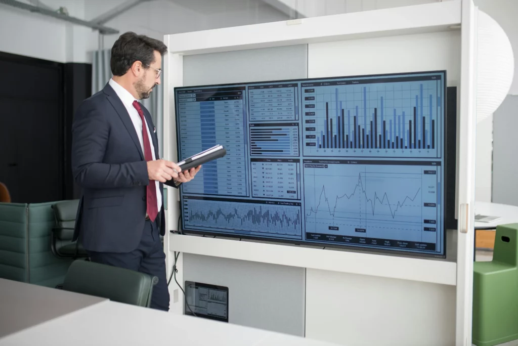 Un homme d'affaires examine des données sur un écran à côté de lui tout en tenant une tablette, dans un bureau moderne.