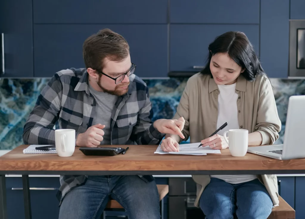 Couple assis à une table remplissant des documents avec une calculatrice et un ordinateur portable.