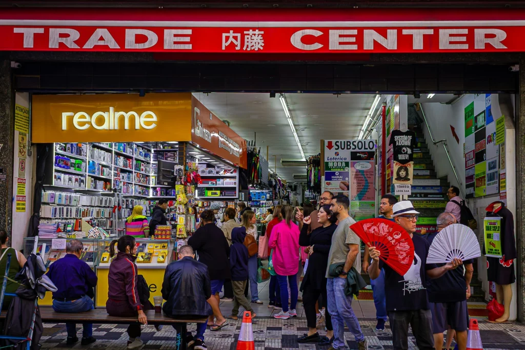 Des gens se promènent et font du shopping devant un centre commercial, avec une vitrine affichant des produits électroniques, dont des téléphones, et un logo "realme".