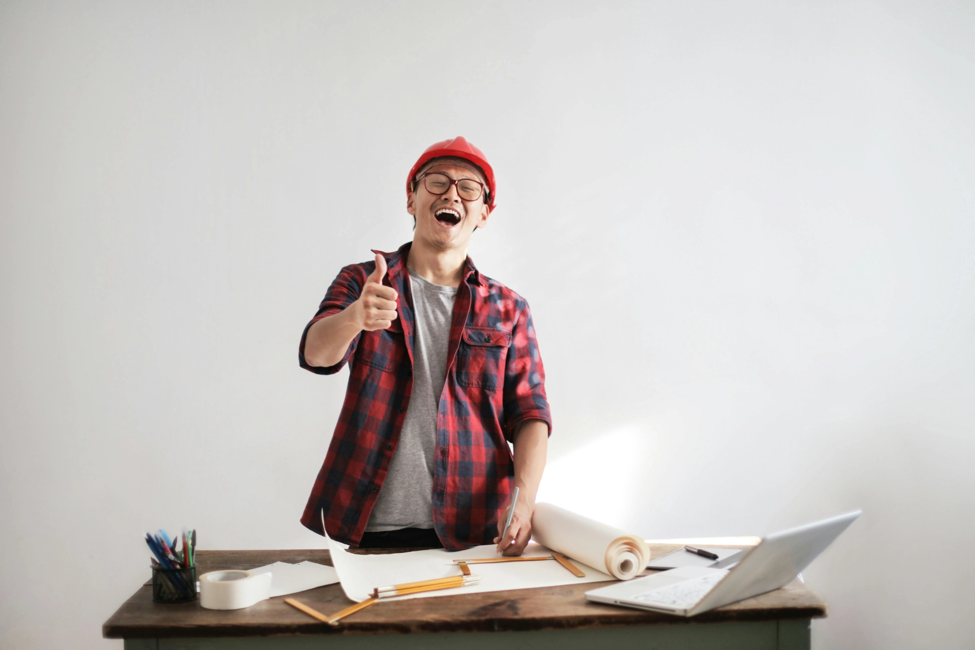 Homme souriant avec un casque de chantier levant le pouce devant un bureau de travail.