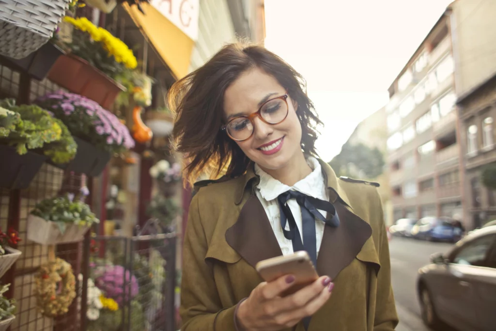 Femme souriante portant des lunettes et consultant son smartphone en marchant dans une rue bordée de fleurs.