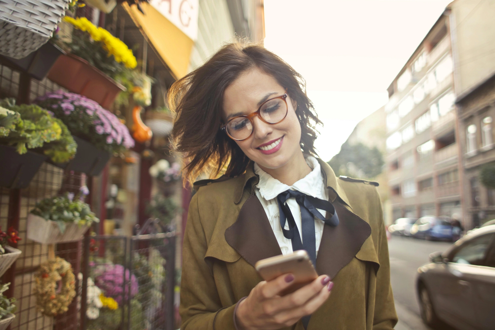 Femme souriante portant des lunettes et consultant son smartphone en marchant dans une rue bordée de fleurs.
