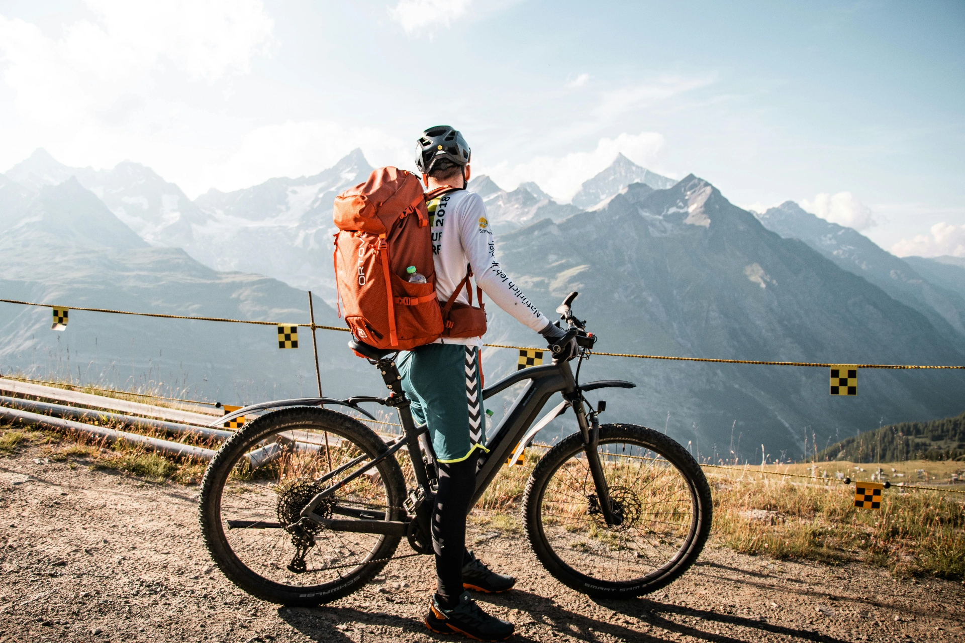 Cycliste avec un sac à dos orange et un VTT contemplant un paysage de montagnes enneigées.

