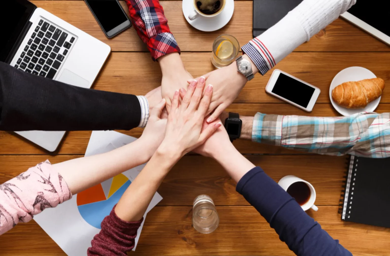 Six personnes joignent leurs mains dans un geste de travail d'équipe autour d'une table en bois dans une agence de conseil en ligne en Belgique, entourées d'ordinateurs portables, de smartphones, de documents, de café et d'un croissant.
