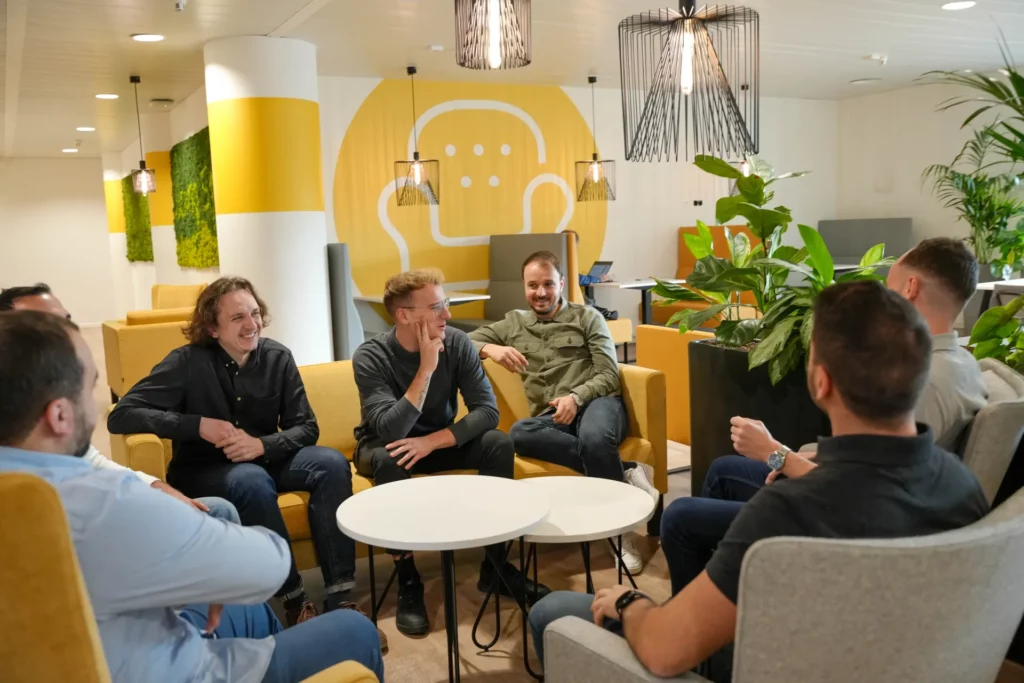 Un groupe de sept hommes est assis dans un salon de bureau moderne, discutant et riant autour de tables blanches, de chaises jaunes et d'œuvres d'art murales en arrière-plan.