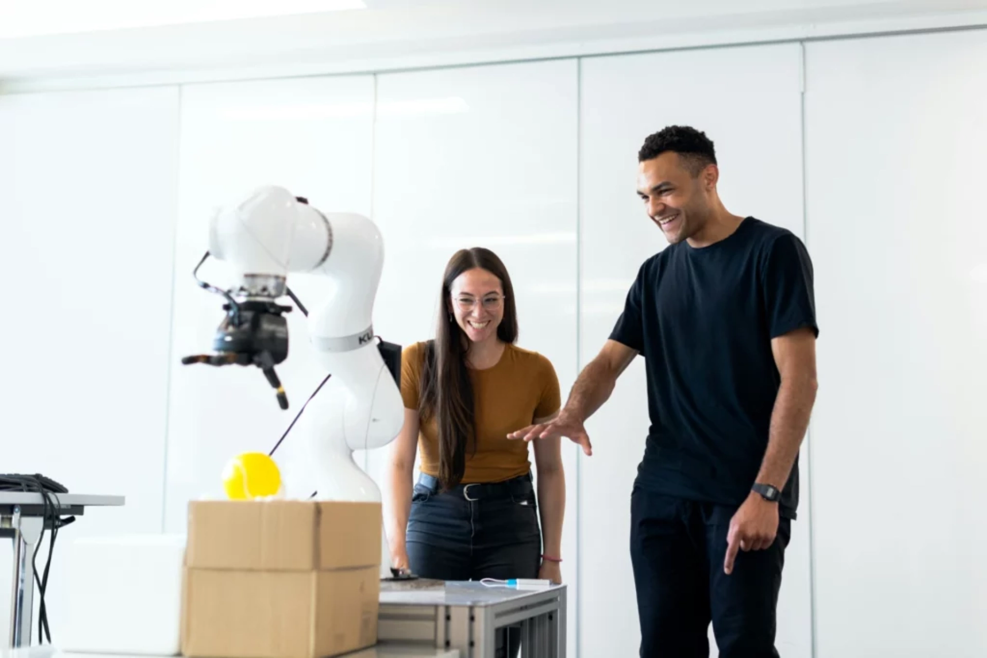 Une femme et un homme interagissent avec un robot dans un laboratoire moderne, observant son action avec enthousiasme. Le robot est équipé d'un bras mécanique, et un objet jaune est posé sur la table devant eux.