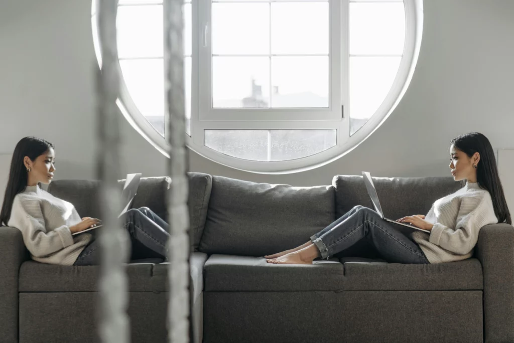 Une jeune femme assise sur un canapé, utilisant un ordinateur portable, avec une grande fenêtre circulaire en arrière-plan, créant une ambiance lumineuse et tranquille.