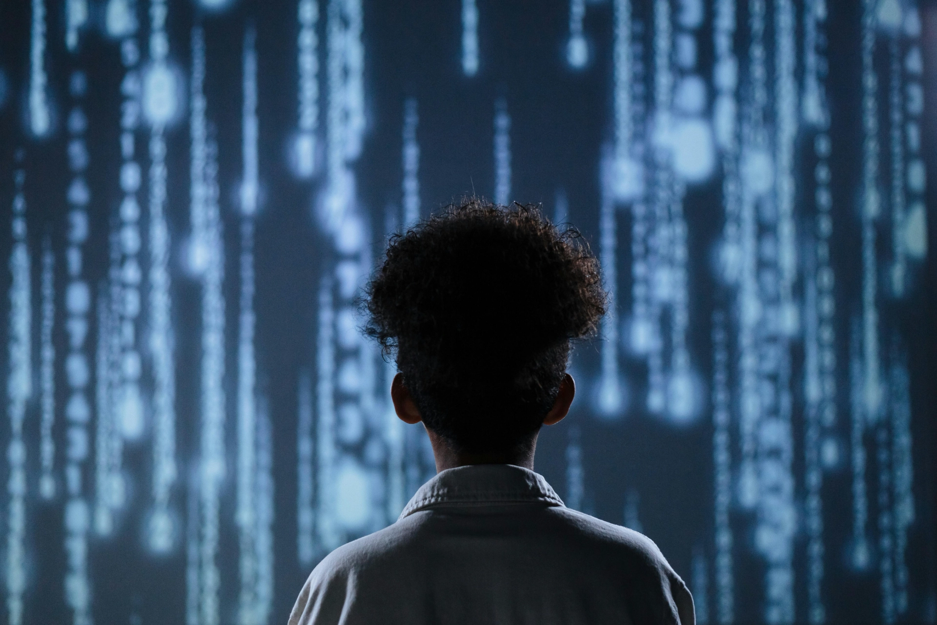 Un enfant regarde un écran projetant des chaînes de chiffres et des symboles, représentant un environnement numérique ou technologique futuriste.