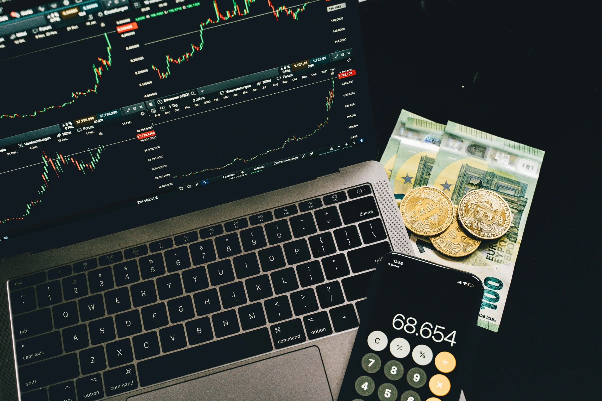 Un ordinateur portable affichant des graphiques boursiers, avec de la monnaie physique et des bitcoins posés à côté, ainsi qu'un téléphone affichant un calcul.