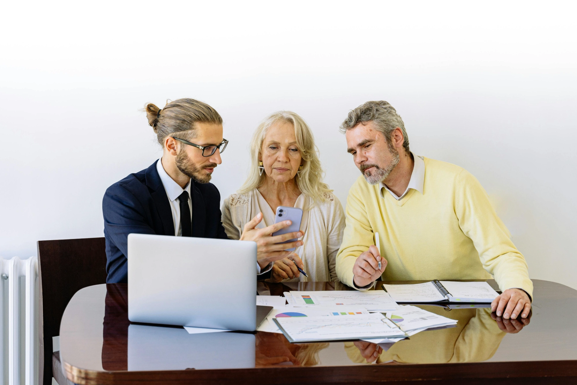 Un homme en costume, une femme âgée et un homme plus âgé, tous trois regardant un téléphone portable en discutant, autour d'une table avec des documents et un ordinateur portable.